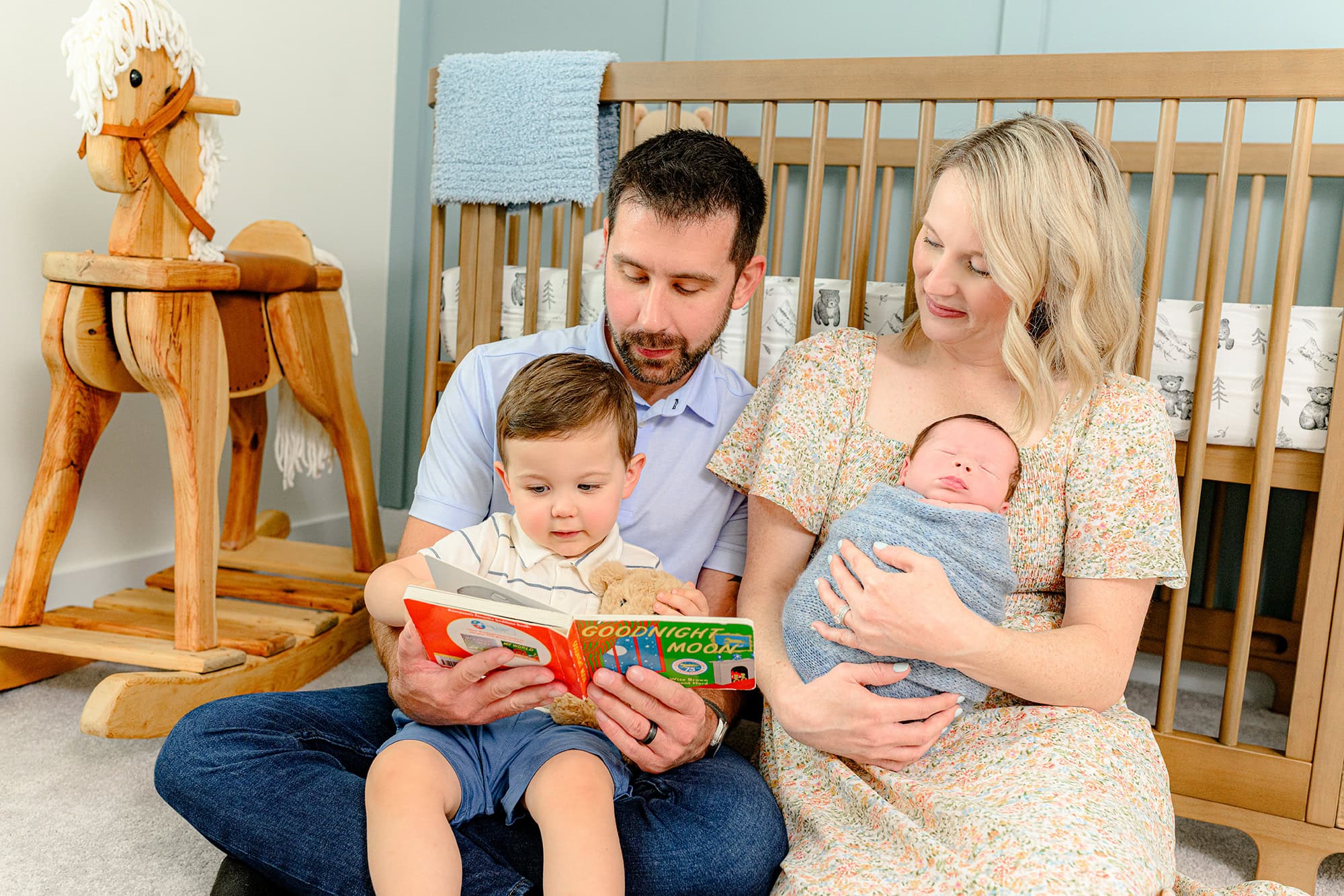 in home lifestyle newborn photo of toddler sibling holding book during baby photos with family with mom holding baby boy