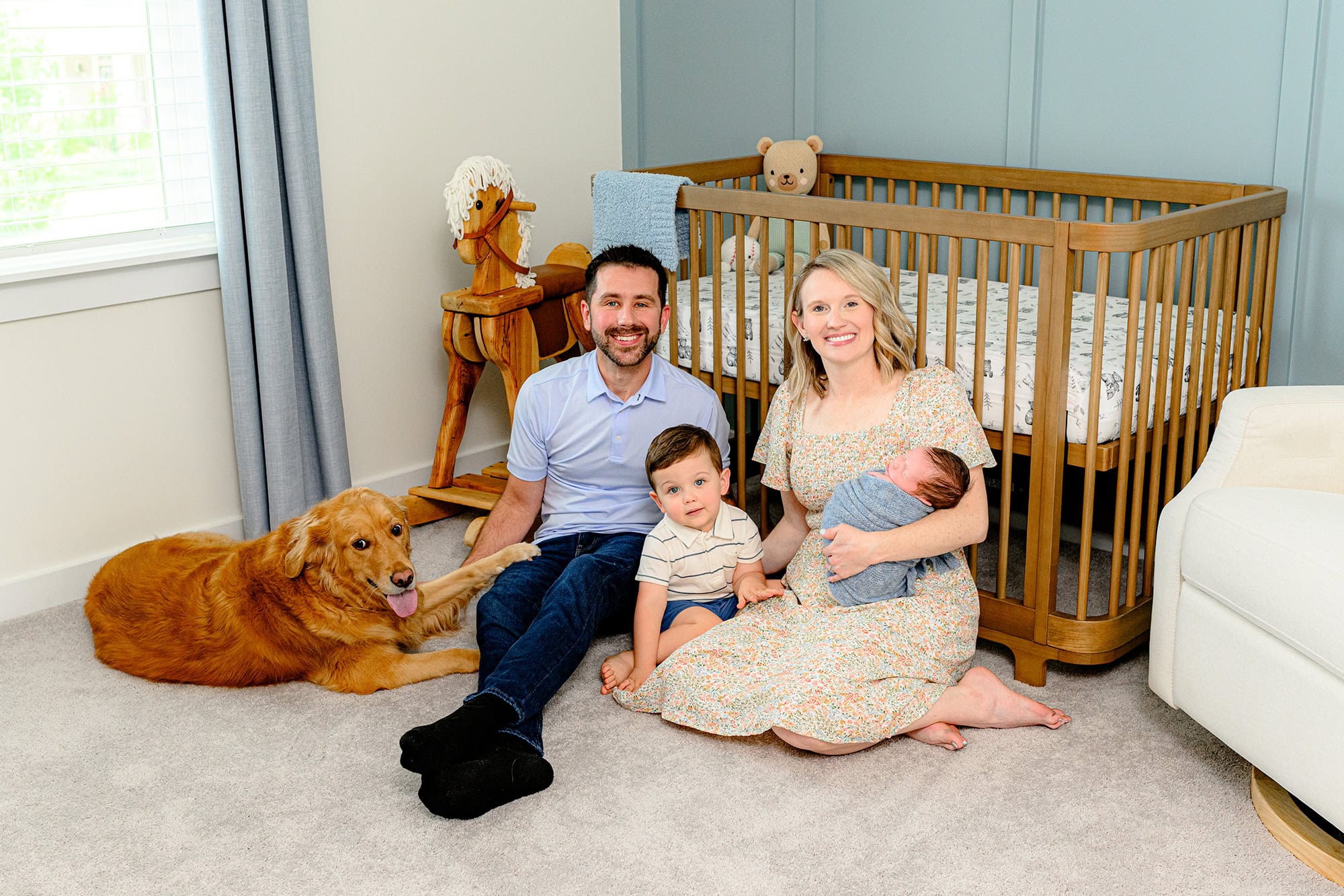 family of four with mom, dad, and toddler sibling sitting on floor of newborns nursery during lifestyle newborn session with family dog included