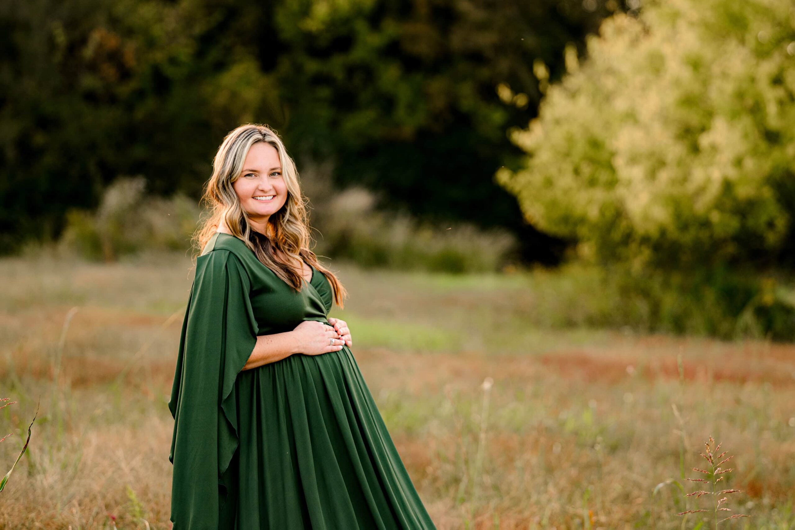 mom to be holding her baby bump standing in tall grasses in a green dress