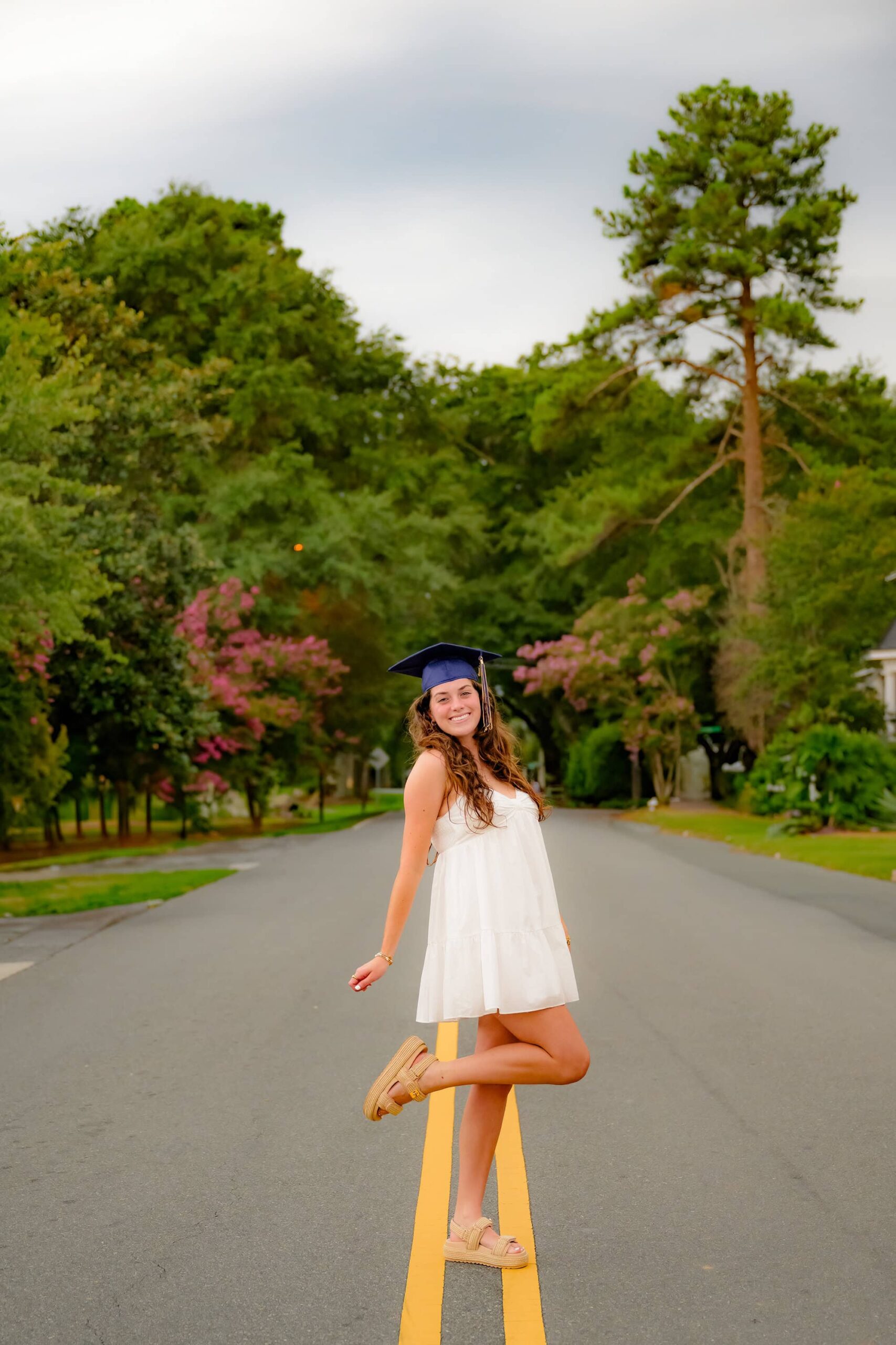 Emma Hines, a Cuthbertson High School senior, strikes a fun and playful pose while wearing her graduation cap in the middle of a downtown Waxhaw street, lifting one foot up while radiating confidence and joy during her senior session.