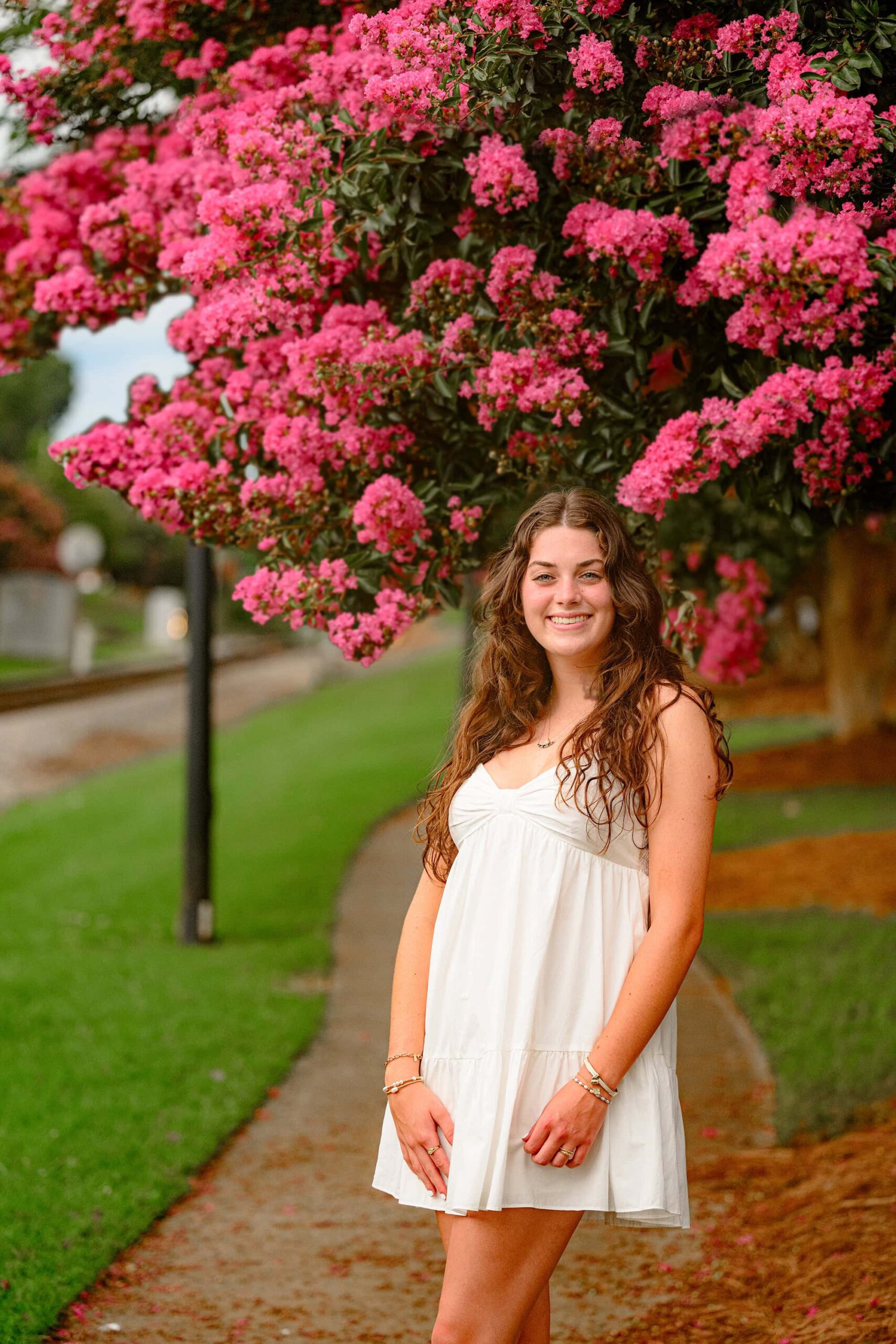 Emma Hines, a green-eyed senior from Cuthbertson High School, stands among vibrant pink crape myrtle blooms in downtown Waxhaw, wearing a flowing white babydoll dress in this soft and dreamy half-body senior portrait.