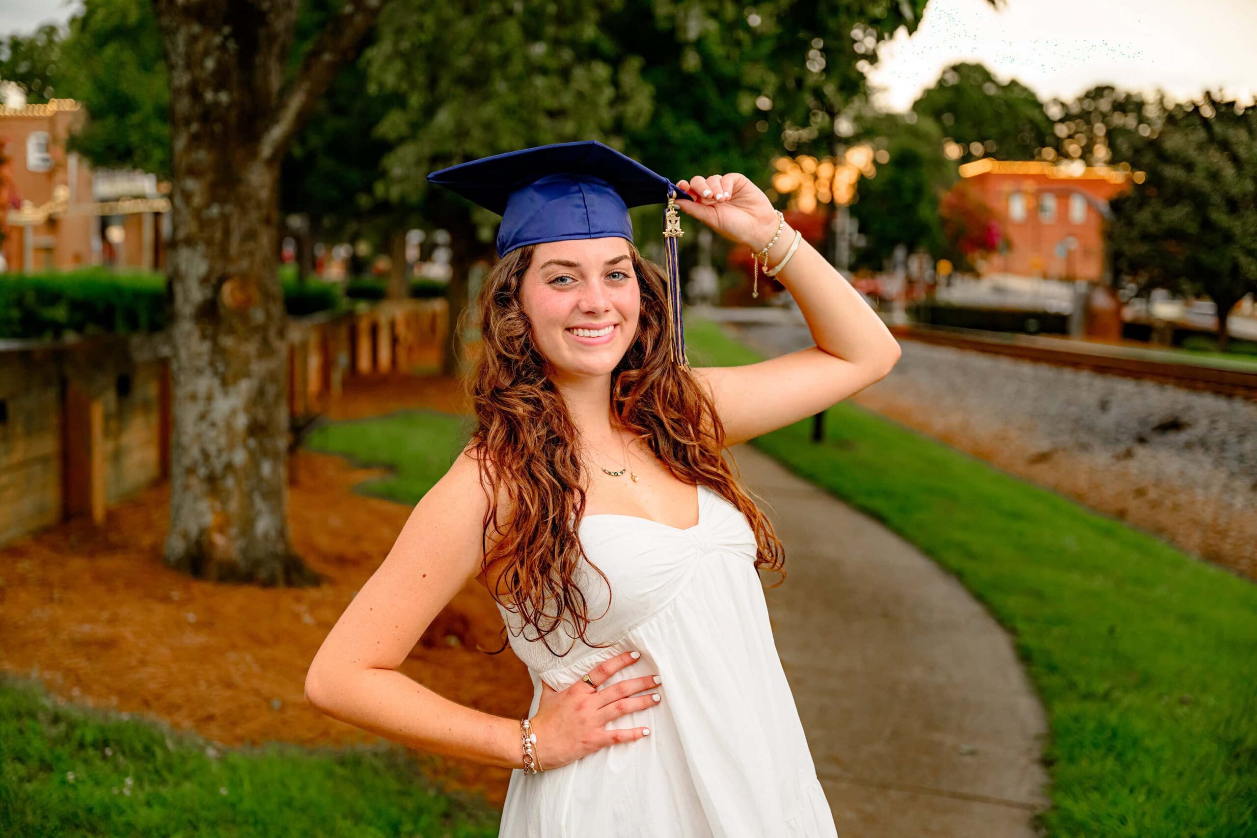 Wearing her blue graduation cap but not the gown, Emma Hines poses elegantly in her dress, gently holding the edge of the cap in her hand, her expression reflecting pride and anticipation for the future.